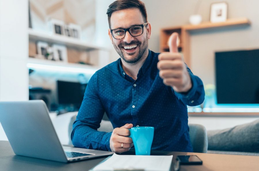 Na obrázku muž v brýlích sedí s kávou u notebooku, kouká do objektivu a s úsměvě ukazuje zvednut palec