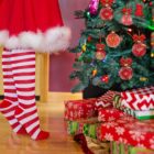 Na obrázku je žena ve vánoční sukni a punčocháčích, jak zdobí vánoční stromek pod kterým leží dárky