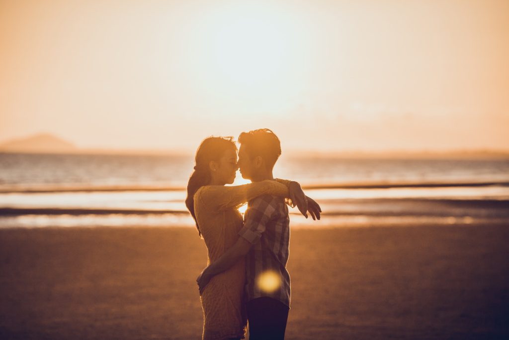 Na obrázku je pár u moře na pláži, objímající se při západu slunce