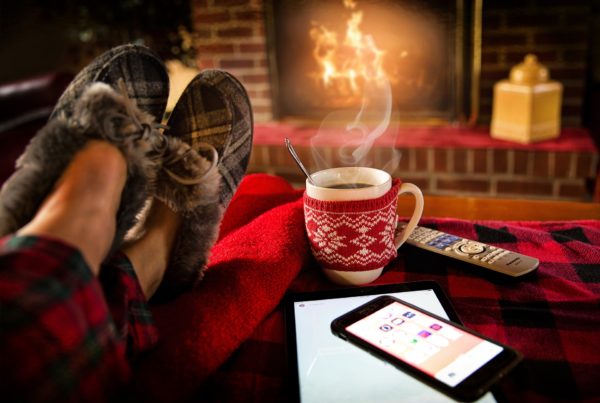 Na obrázku jsou ve Vánoční pohodě překřížené nohy v papučích, zapálený krb a sváteční hrnek s horkým kakaem