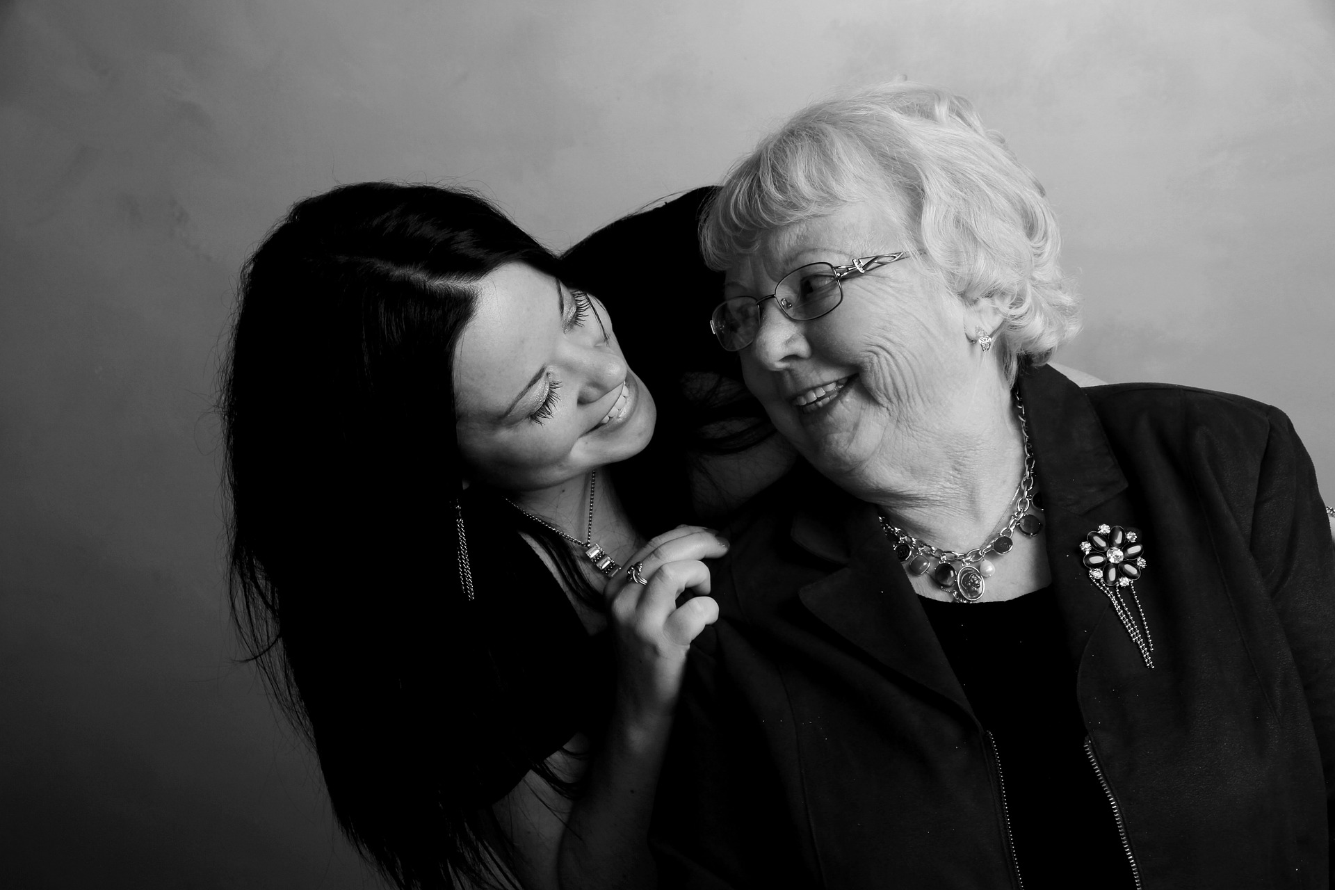 Na černobílém obrázku je žena stojící za starší ženou, ke které se ohýbá a společně se na sebe usmívají