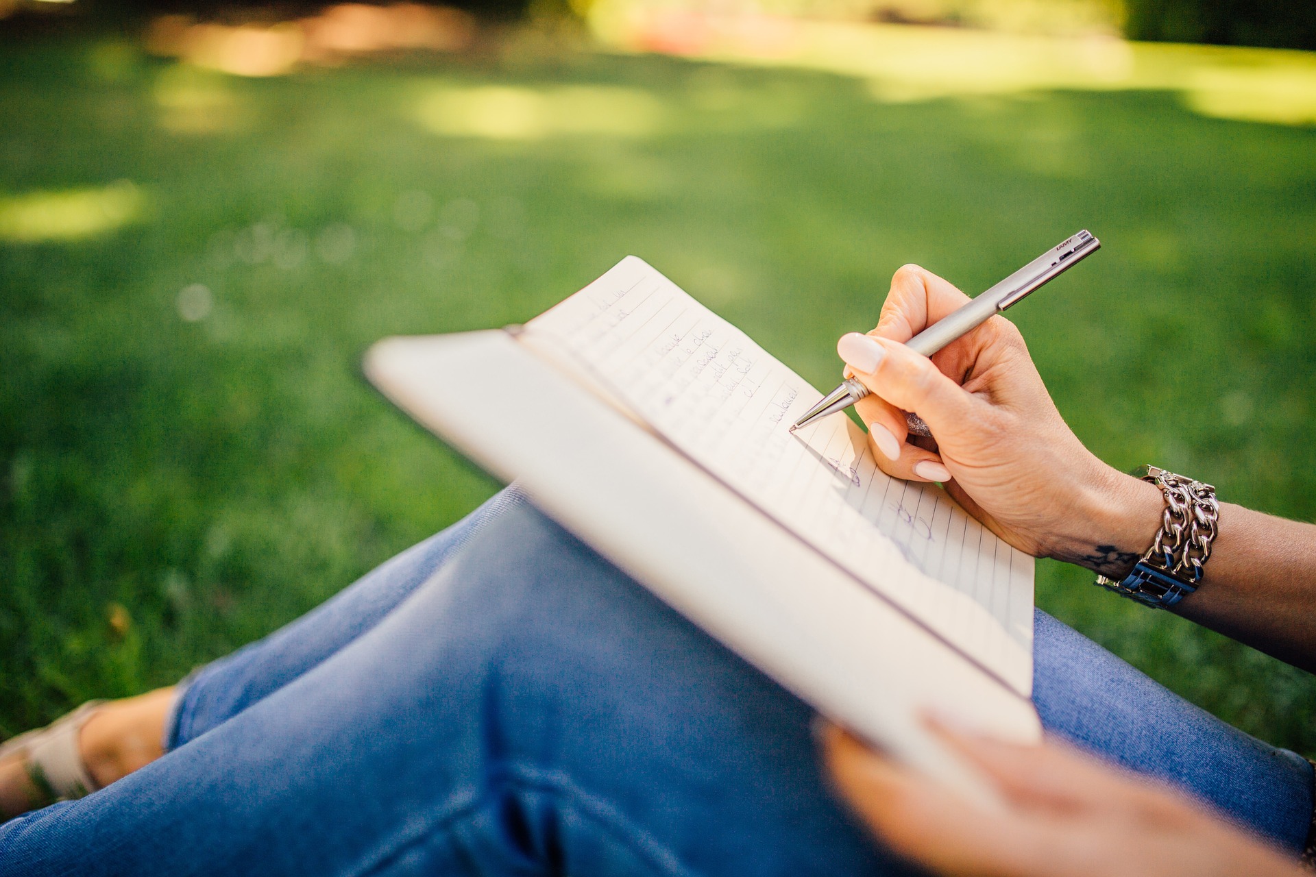 Na obrázku je žena sedící v trávě, která si zapisuje do deníku