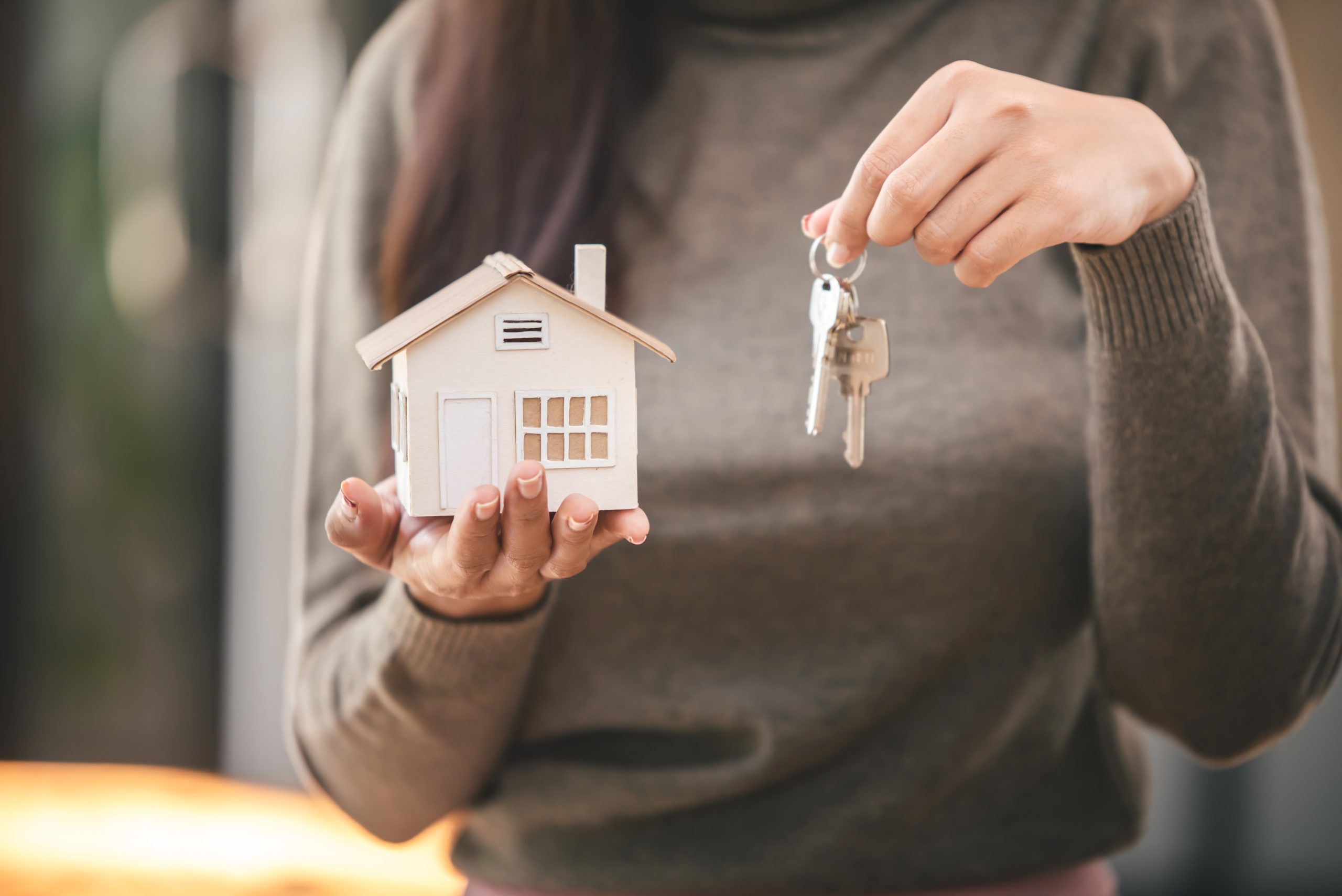 Na obrázku je žena, držící v jedné ruce model domku a v druhé ruce klíče od dveří
