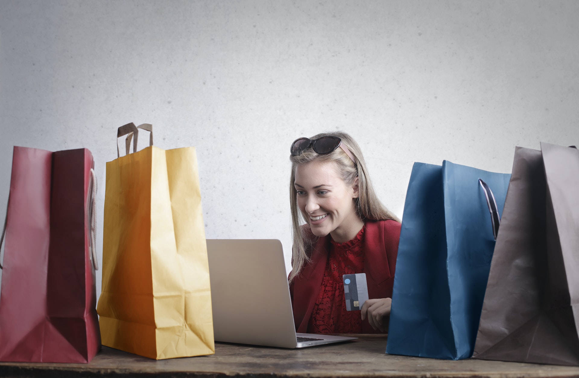 Na obrázku je žena s notebookem držící platební kartu, obklopená nákupními taškami