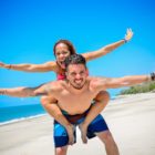 Na obrázku je na pláži muž se smějící se ženou, která mu sedí na zádech s rozpřaženýma rukama