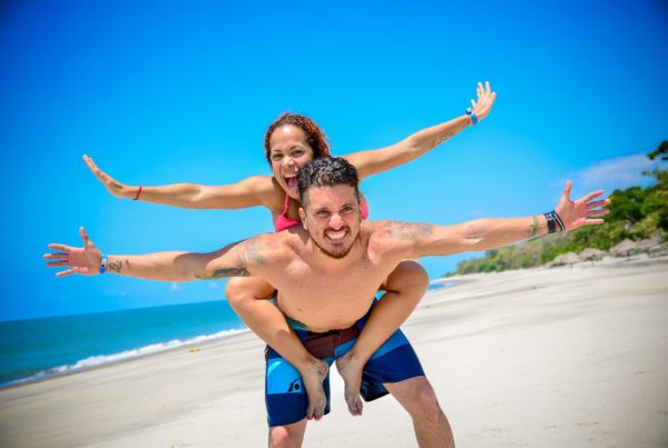 Na obrázku je na pláži muž se smějící se ženou, která mu sedí na zádech s rozpřaženýma rukama