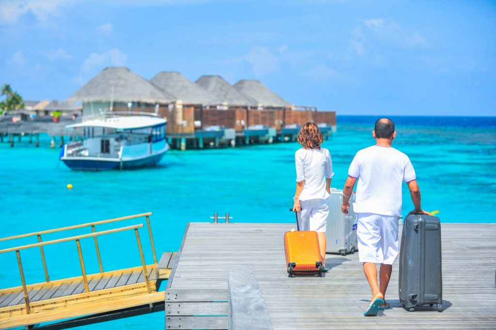 Na obrázku je muž a žena s kufry u azurově modrého moře s chatkami