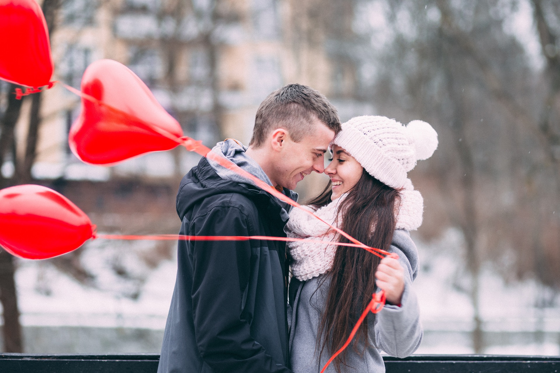 Na obrázku je zamilovaný pár v zimě venku, žena v ruce drží tři nafukovací srdíčka.