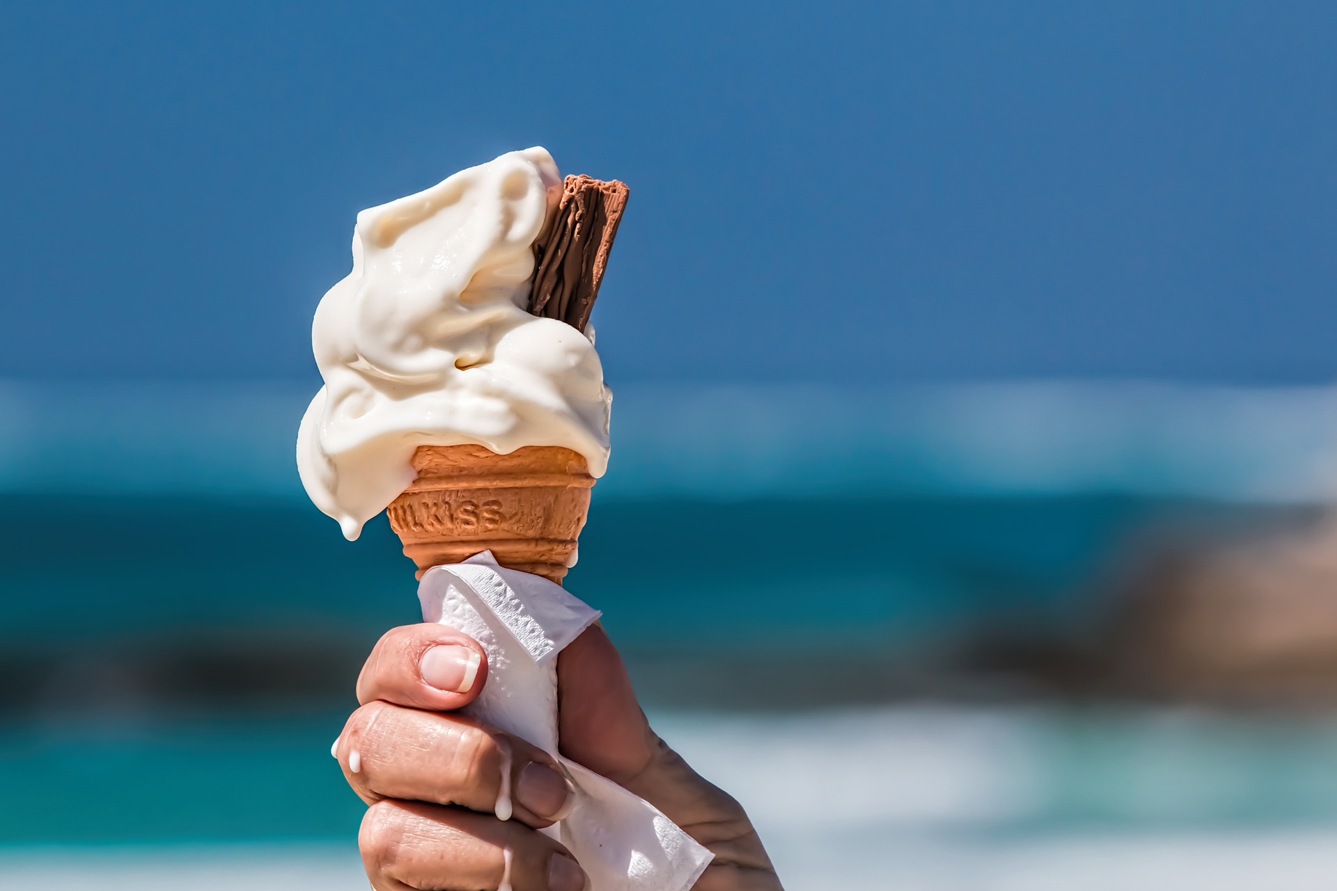 Na obrázku je ruka držící kornoutek se zmrzlinou a mořem v pozadí