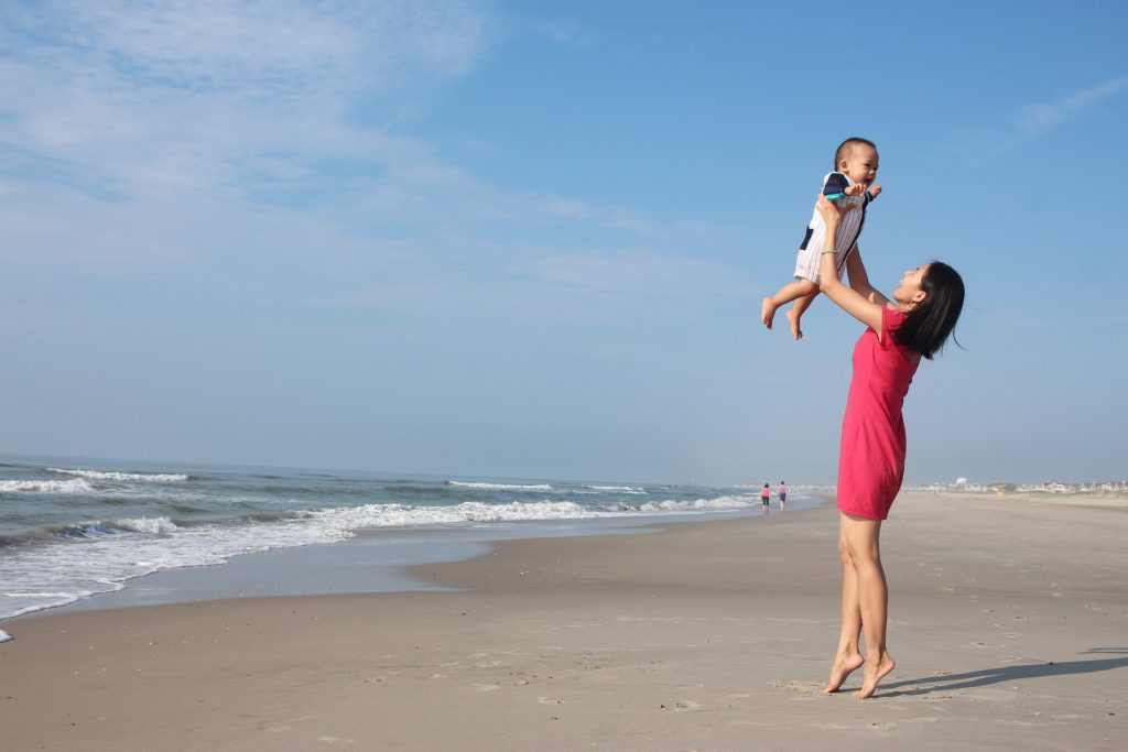 Na obrázku zvedá žena na pláži do vzduchu kojence