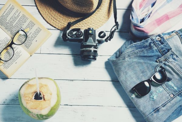 Na obrázku jsou věci na dovolené - oblečení, knížka, brýle, foťák a kokosový ořech s brčkem
