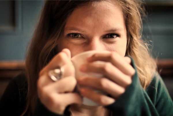 Na obrázku je žena, která pije kávu z hrnku a usmívá se do objektivu