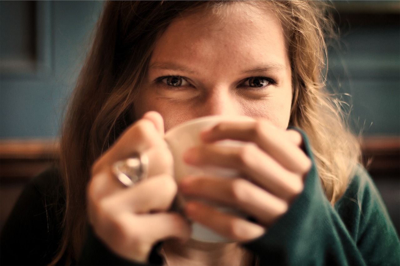 Na obrázku je žena, která pije kávu z hrnku a usmívá se do objektivu